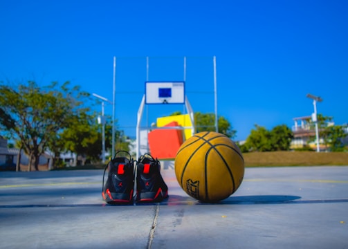 A basketball and a pair of black sports shoes are placed on an outdoor basketball court. A basketball hoop with a backboard is visible in the background, and the court is surrounded by trees and some buildings under a clear blue sky.