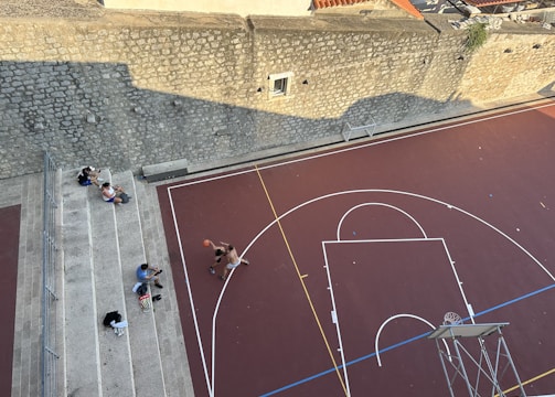 An aerial view of an outdoor basketball court with two people playing basketball. The court has clearly marked lines and a hoop with a backboard. Three individuals are sitting on the bleachers to the side of the court, facing towards the game. The court is bordered by a stone wall with a small window, casting a shadow on part of the court.